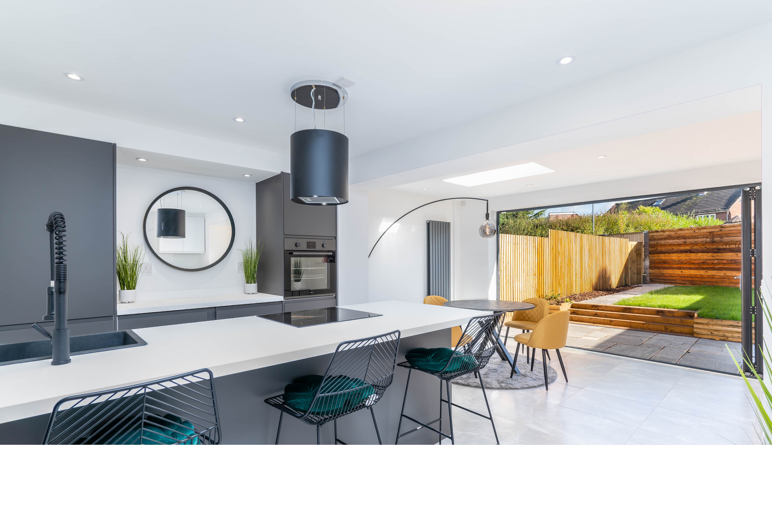 Modern kitchen with white island, black chairs with cushions, gray cabinets, and a view of a fenced backyard with grass, patio, and trees.