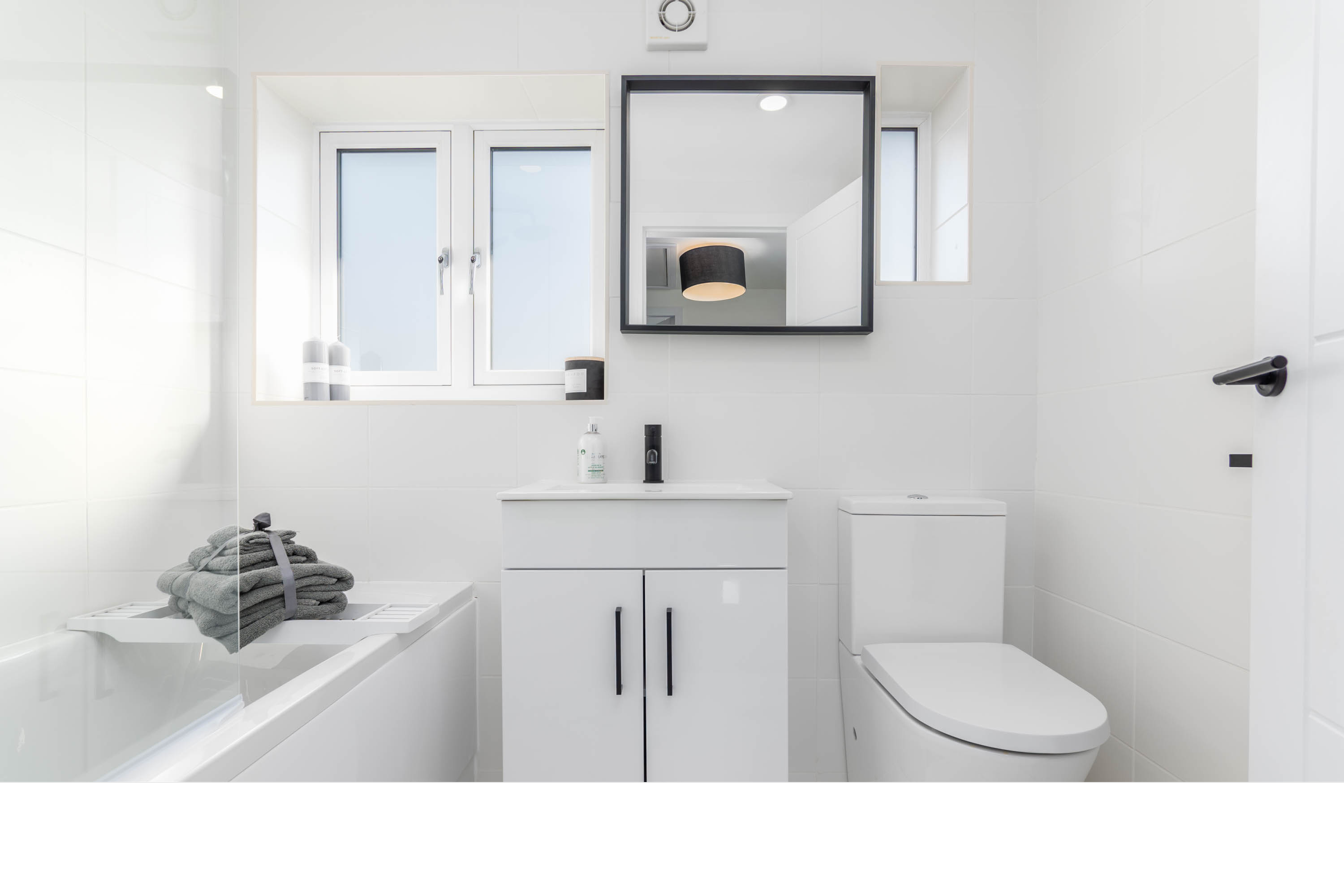 A modern, minimalist bathroom with white walls, a bathtub with stacked gray towels, a small white vanity with a mirror, a window, and a toilet.