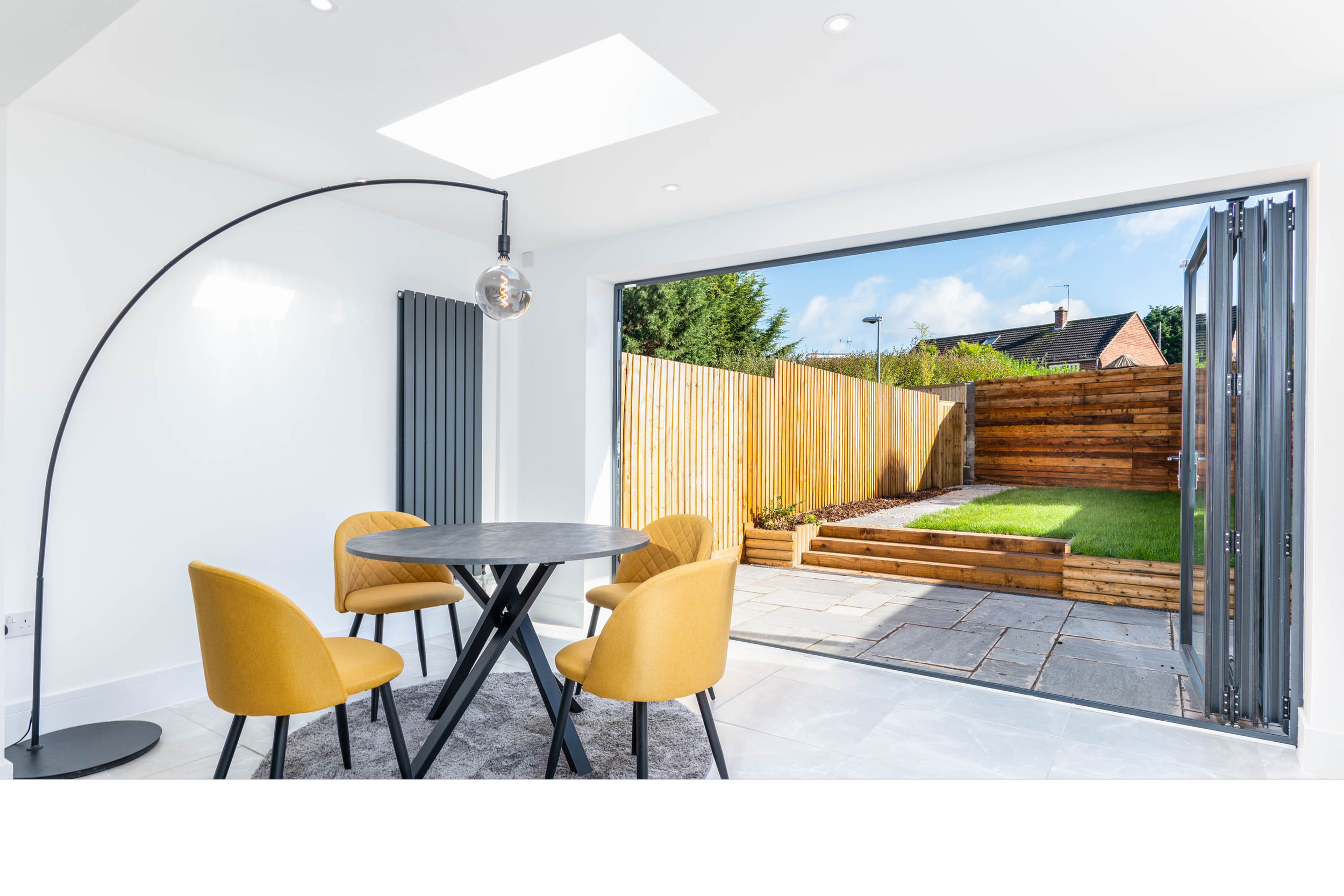 Modern dining area with yellow chairs around a black table, open to a backyard with a wooden fence, patio stones, and a grassy lawn under a blue sky.