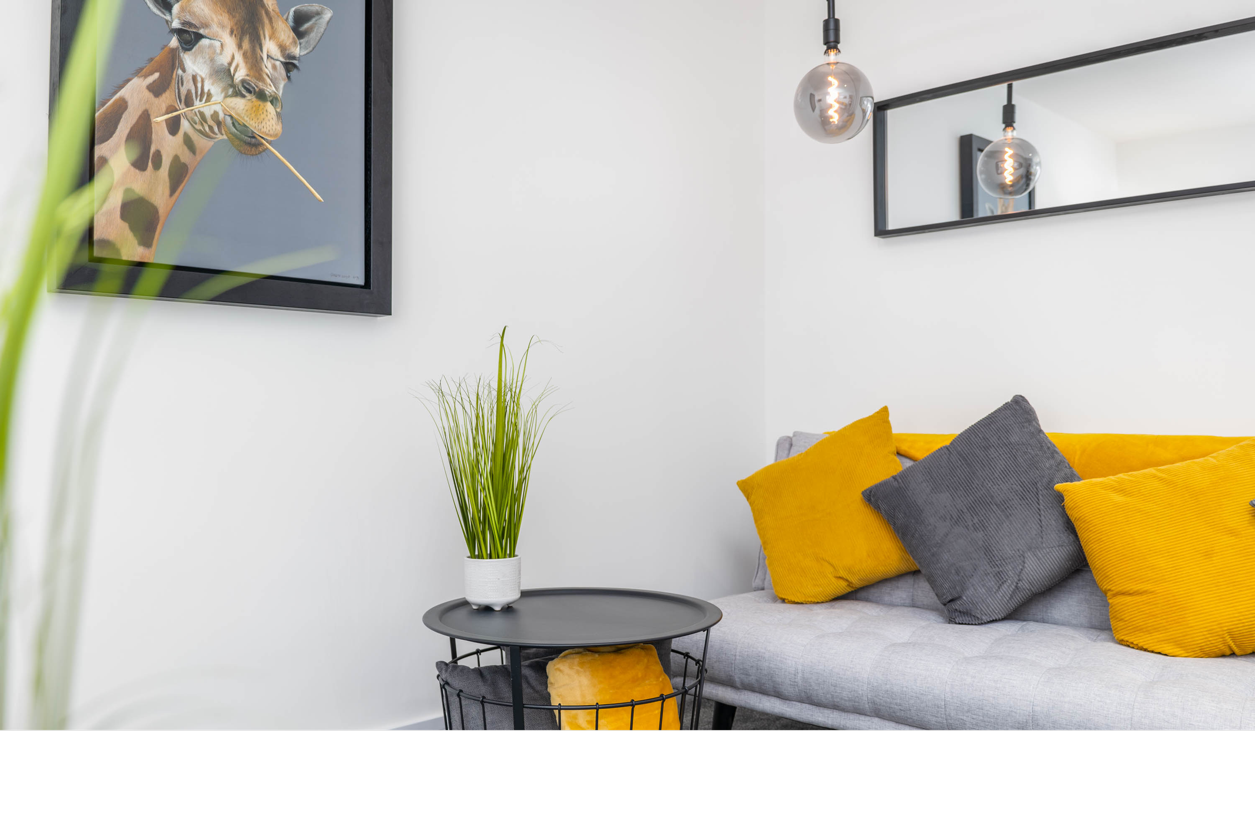 Modern living room with a gray sofa and yellow and gray pillows, a black side table with a green plant, a large wall mirror, a pendant light bulb, and a framed giraffe artwork.