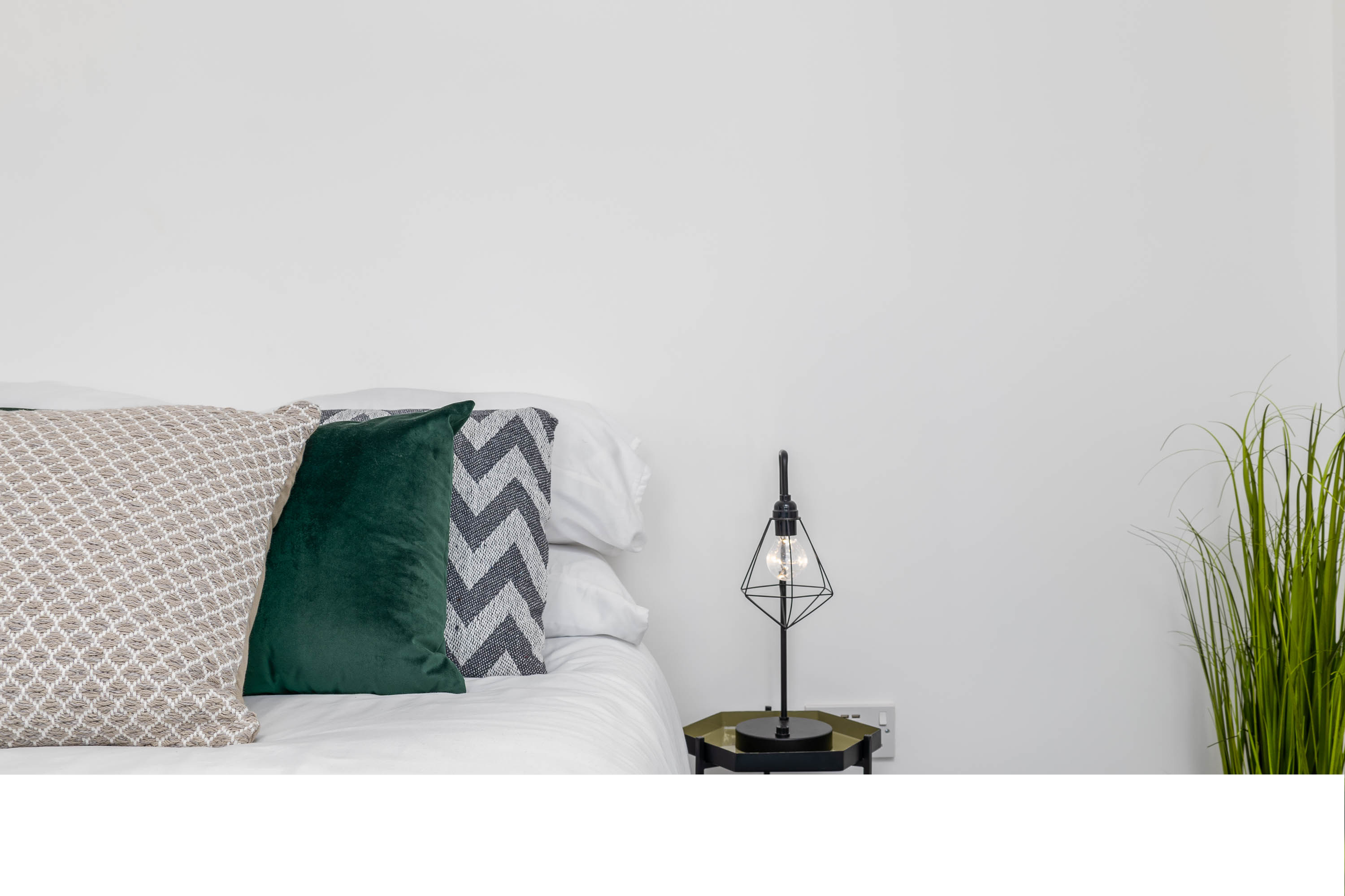 Corner of a bedroom with a bed that has multiple pillows and a modern black side table with a geometric table lamp, against a white wall with a green plant on the right.