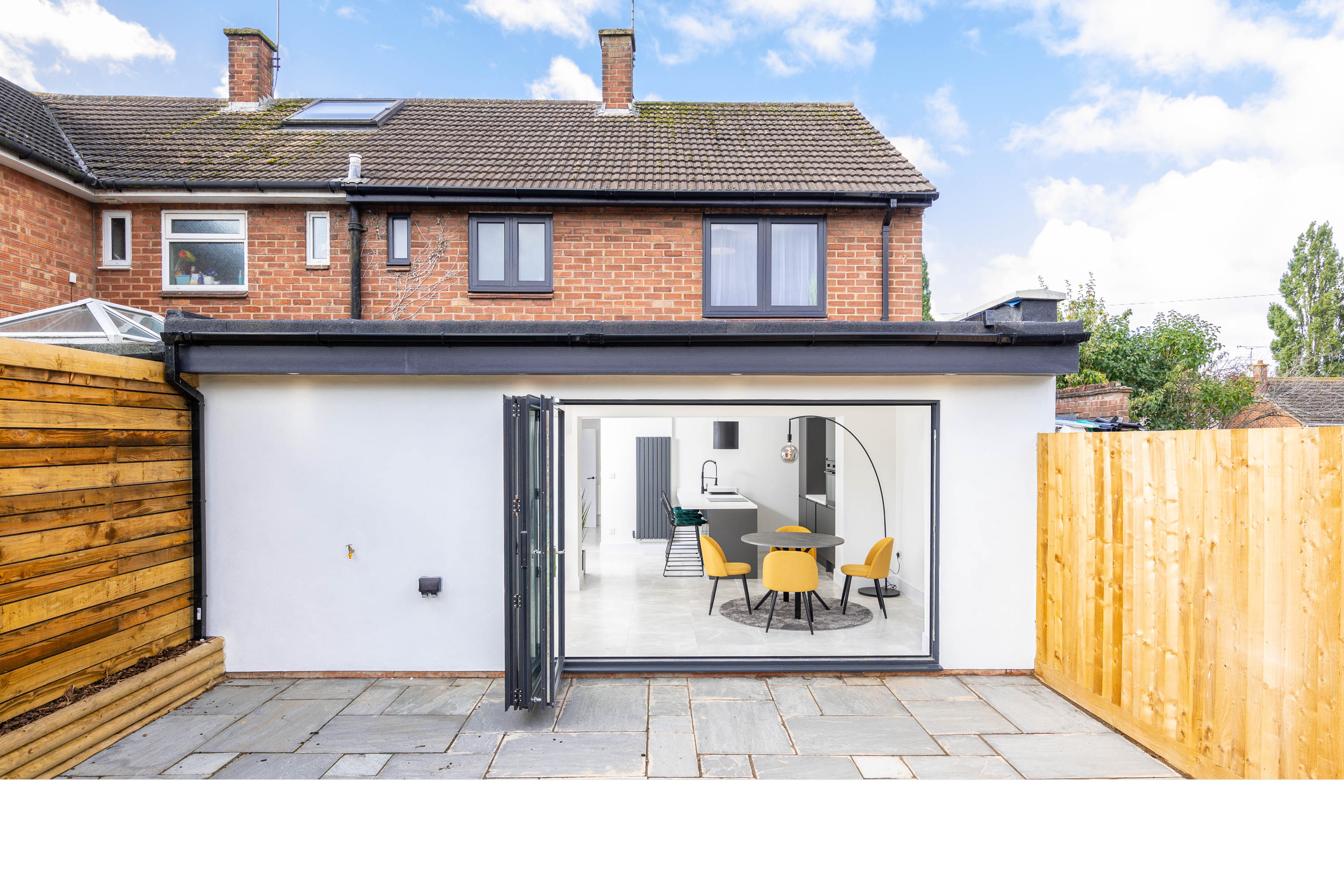 A modern backyard view showing a minimalist white extension with an open folding glass door. Inside, there is a round dining table with four yellow chairs, a small kitchen, and interior decor. The outdoor patio has stone tiles, and the yard is enclos