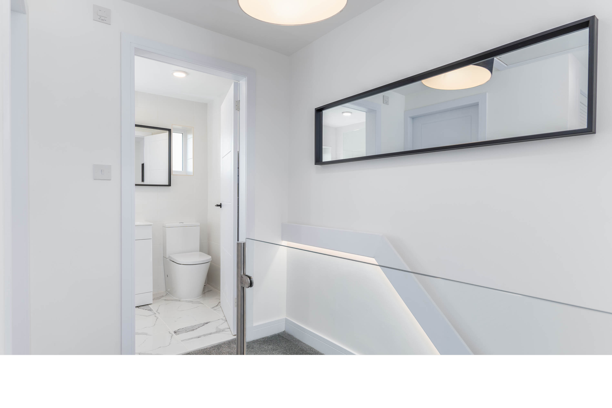 Modern hallway with white walls, a horizontal mirror on the wall, and a staircase with a glass railing. An open door reveals a small bathroom with a toilet, a window, and a white washing machine.