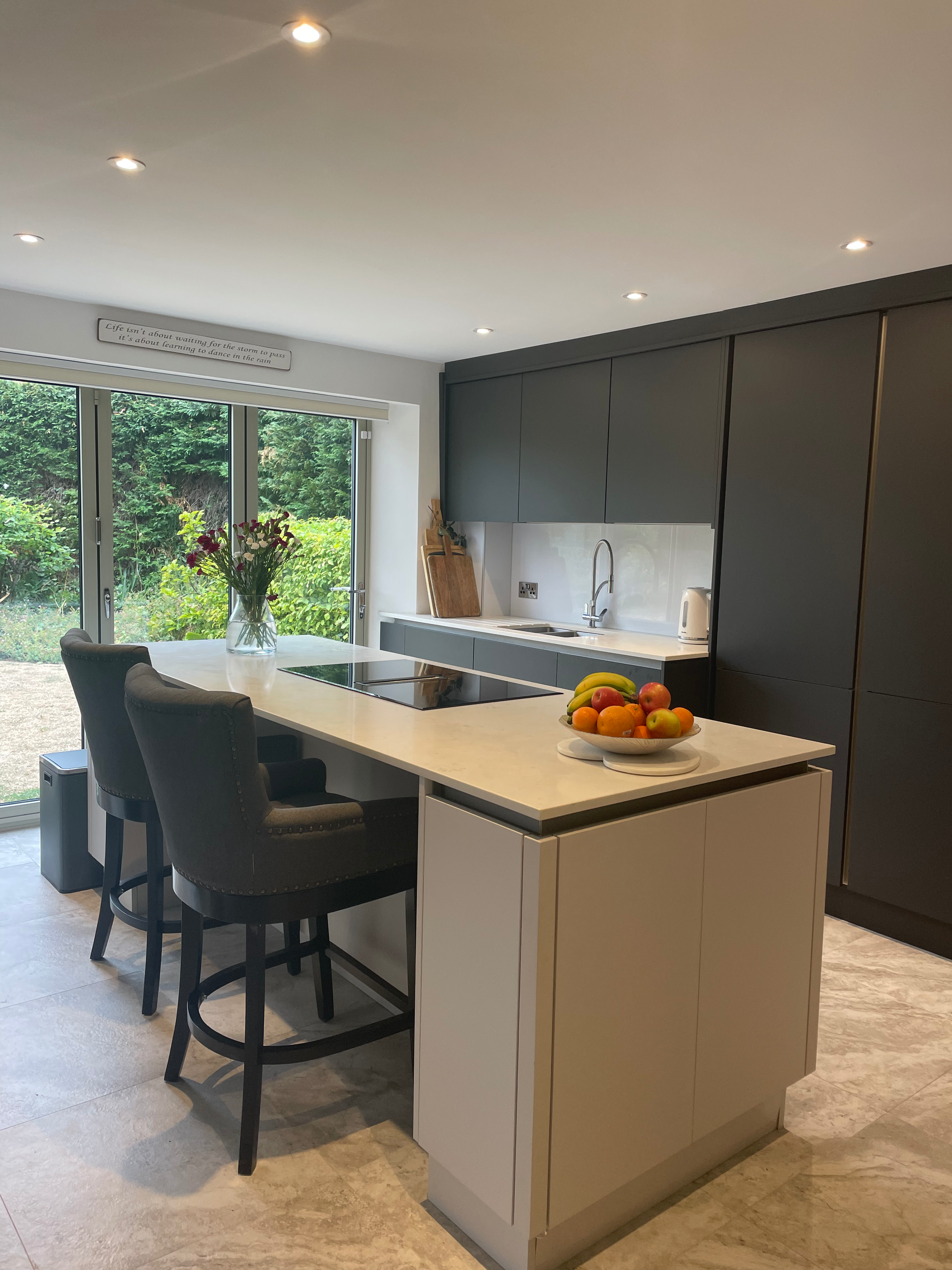 Modern kitchen with an island, bar stools, a bowl of assorted fruit, and sliding glass doors showing a garden outside.