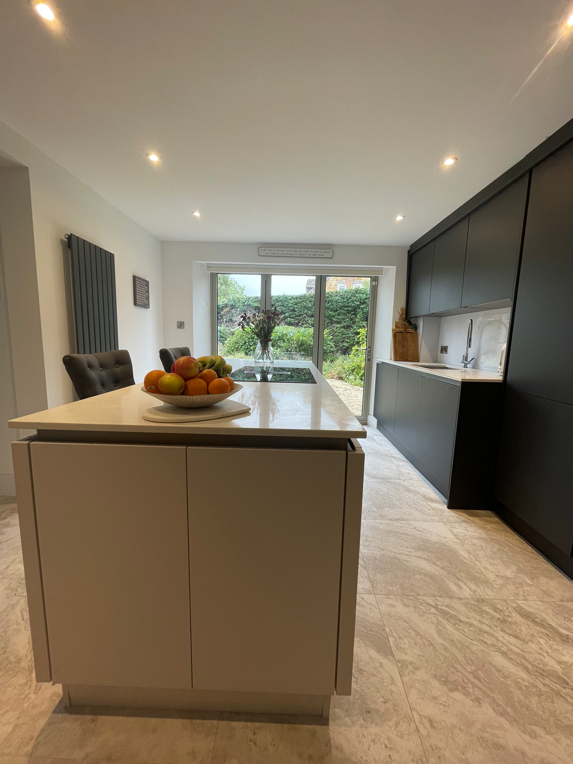 Modern kitchen with beige island counter, fruit bowl, dark cabinets, large window, outdoor garden, and dining area.