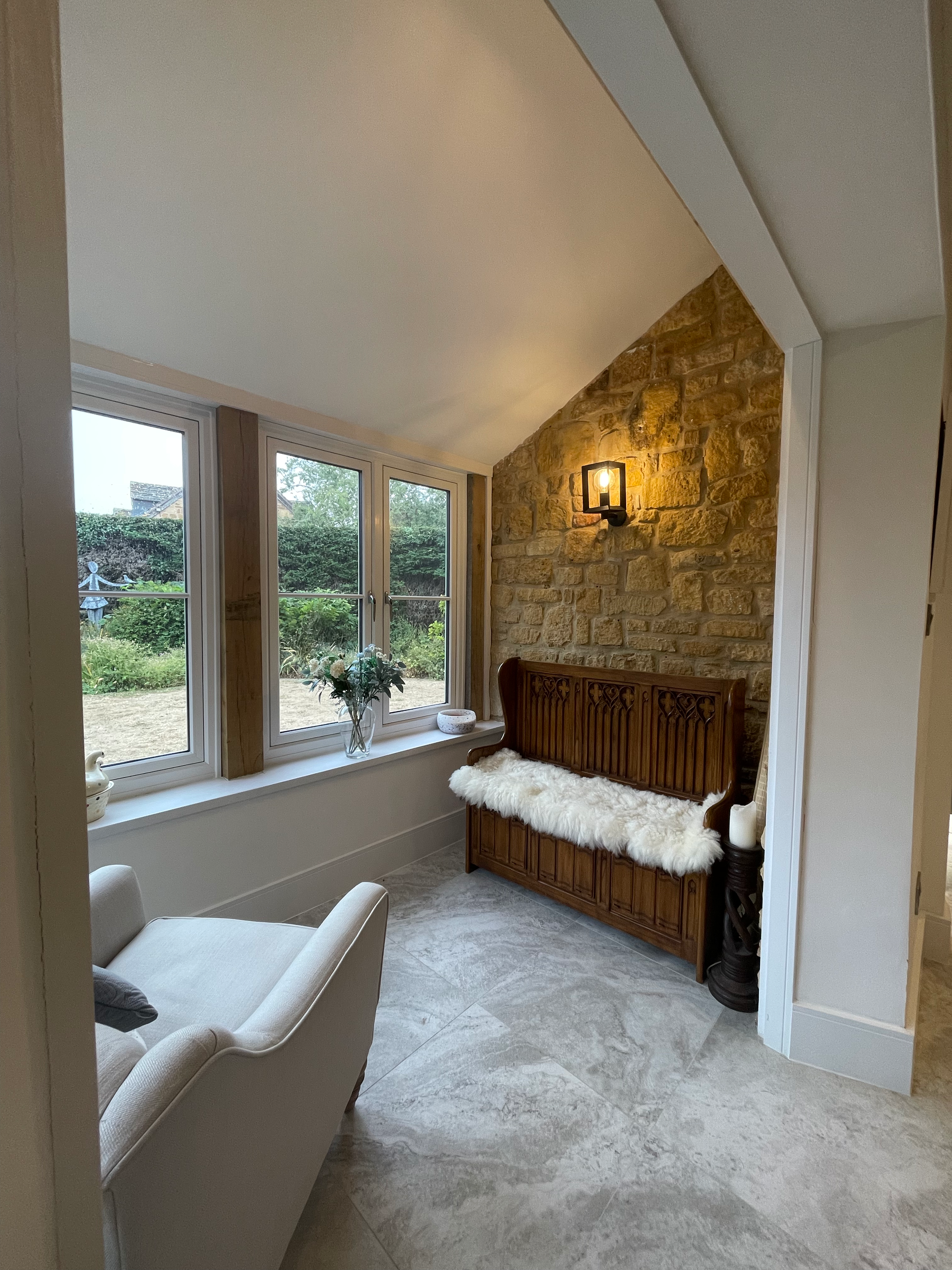 Cozy corner of a living room with large windows, a wooden bench with a white fur throw, a white armchair, and a brick accent wall with a wall-mounted light fixture.