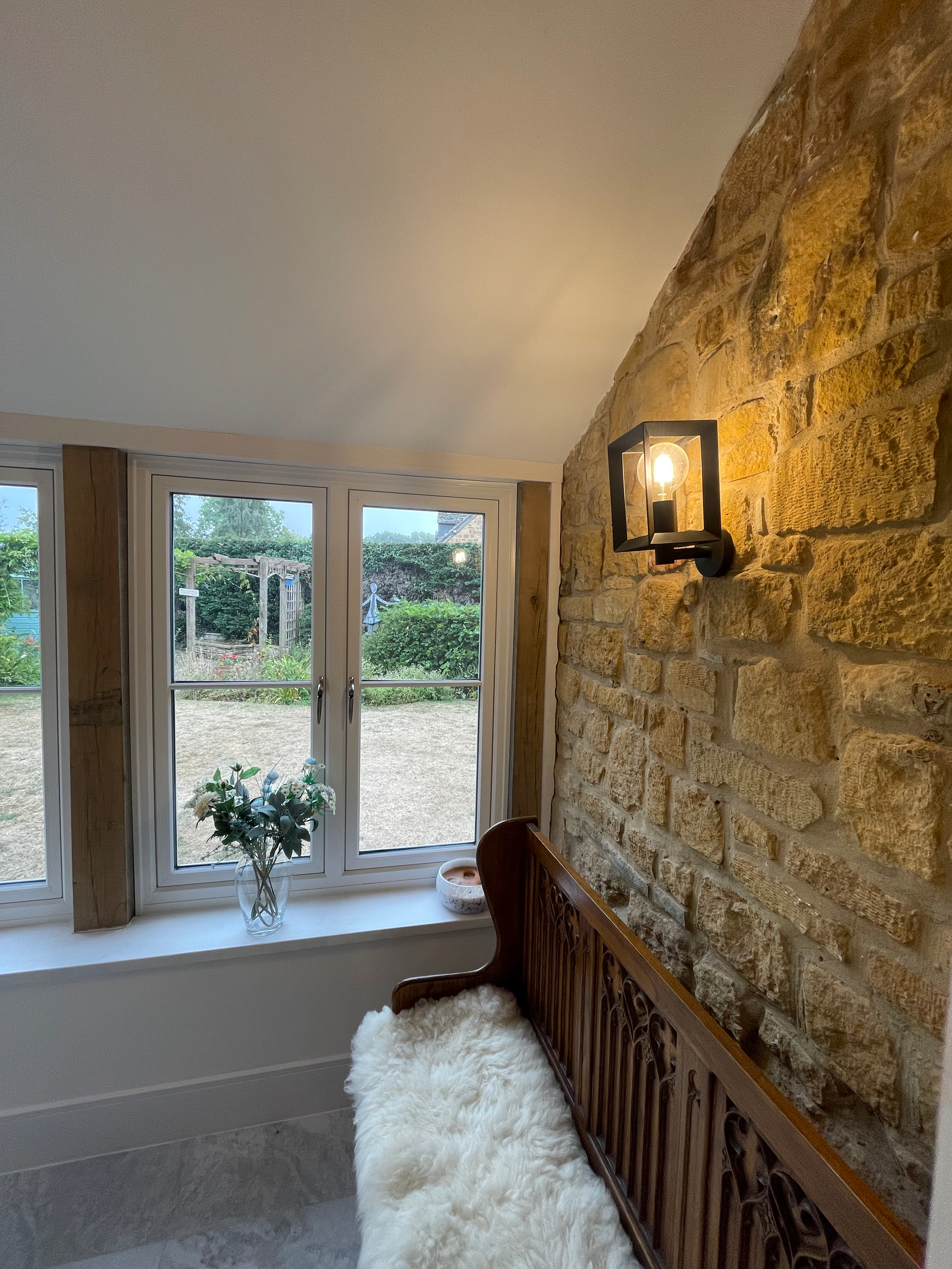 Interior of a cozy room with a stone wall, modern wall sconce, wooden bench with a furry cushion, a window with a view of a garden, a vase of flowers, and a small decorative object on the window sill.