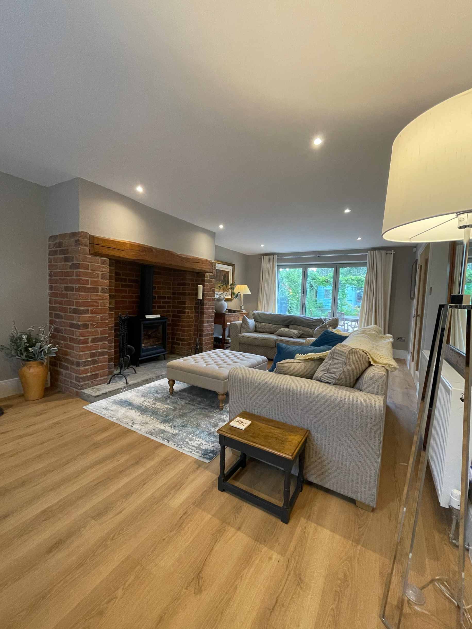 Living room with a brick fireplace, large windows, hardwood flooring, multiple sofas, an ottoman, a side table, and a floor lamp.