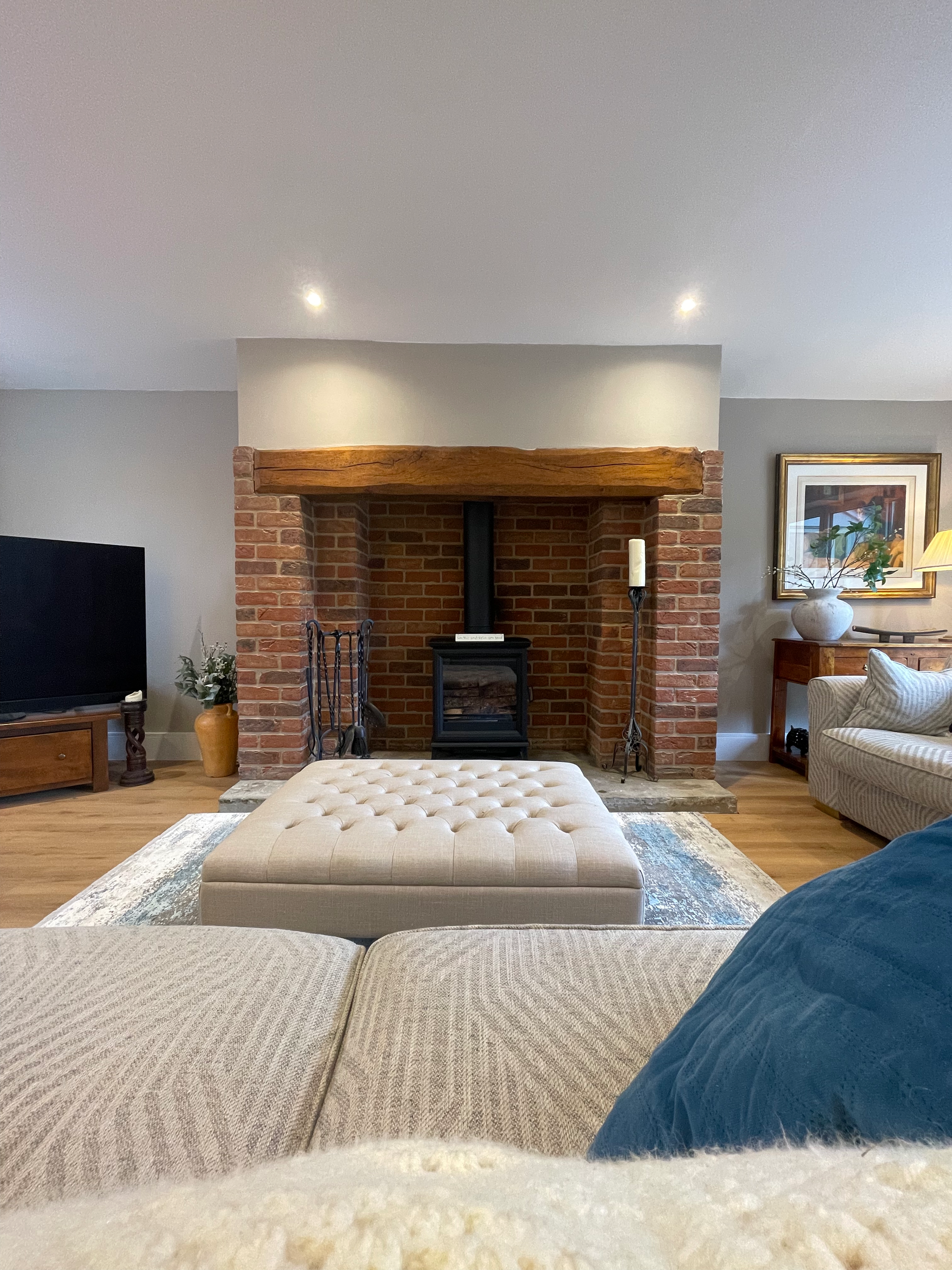 Living room with brick fireplace, beige tufted ottoman, sofa with blue and beige pillows, TV, potted plant, framed artwork, and side table with a lamp.