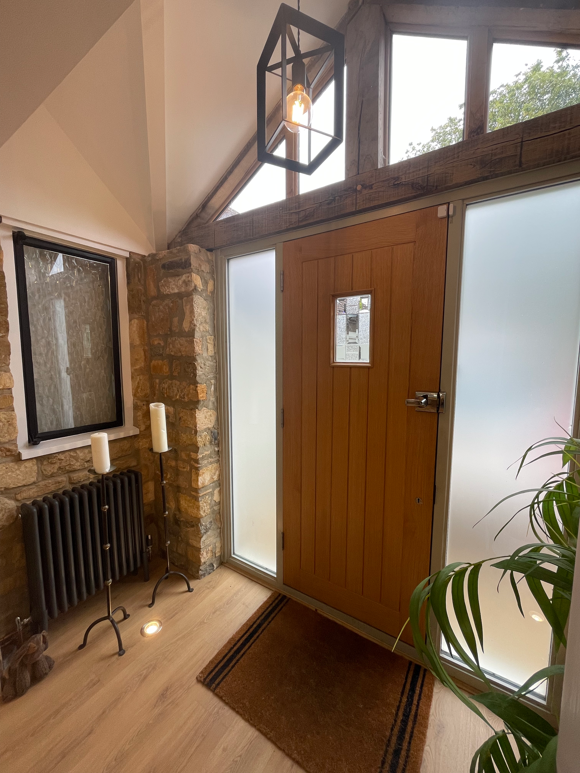Entryway with wooden door and frosted glass side panels, stone wall, black radiator, two candle stands with white candles, a small window, hanging geometric light fixture, wooden floor, and a small rug.