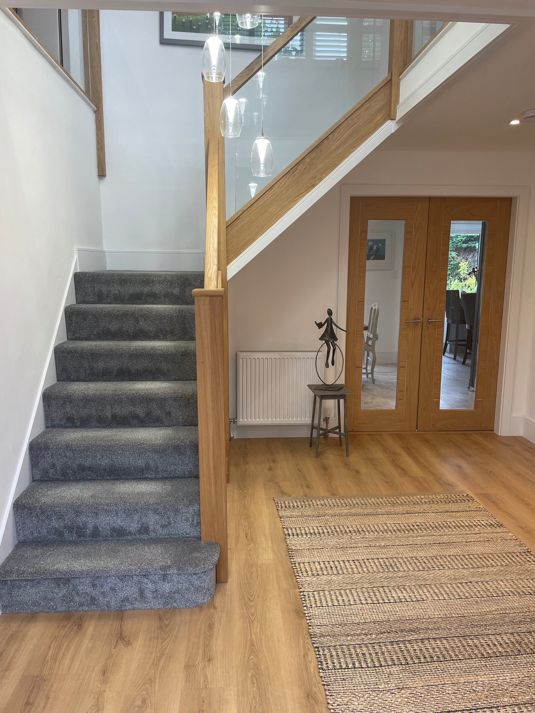 Interior of a house with a staircase covered in gray carpet, wooden handrail, glass paneled wall, two wooden doors with mirrors, a small statue of a dancer, a stool, and a striped rug on a wooden floor.
