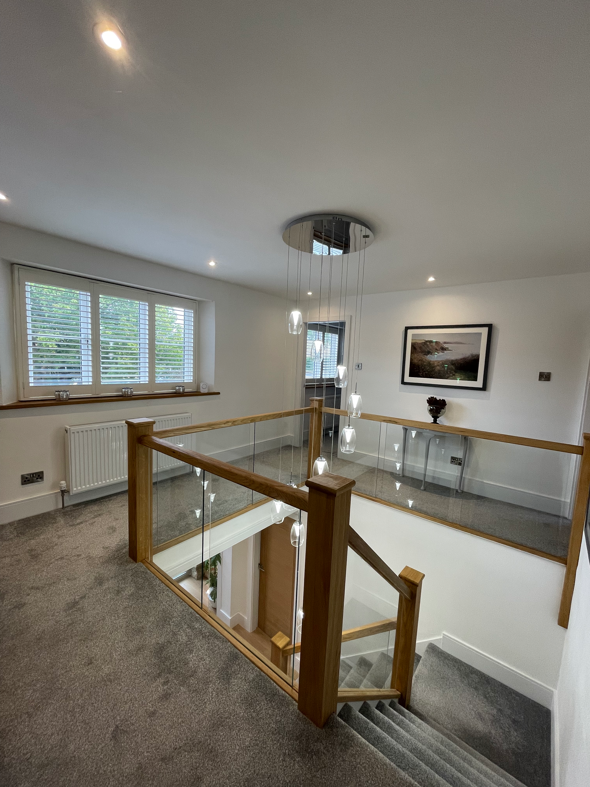 Interior view of a staircase landing with a modern chandelier, framed landscape photo on white wall, window with blinds, and wooden and glass railing.