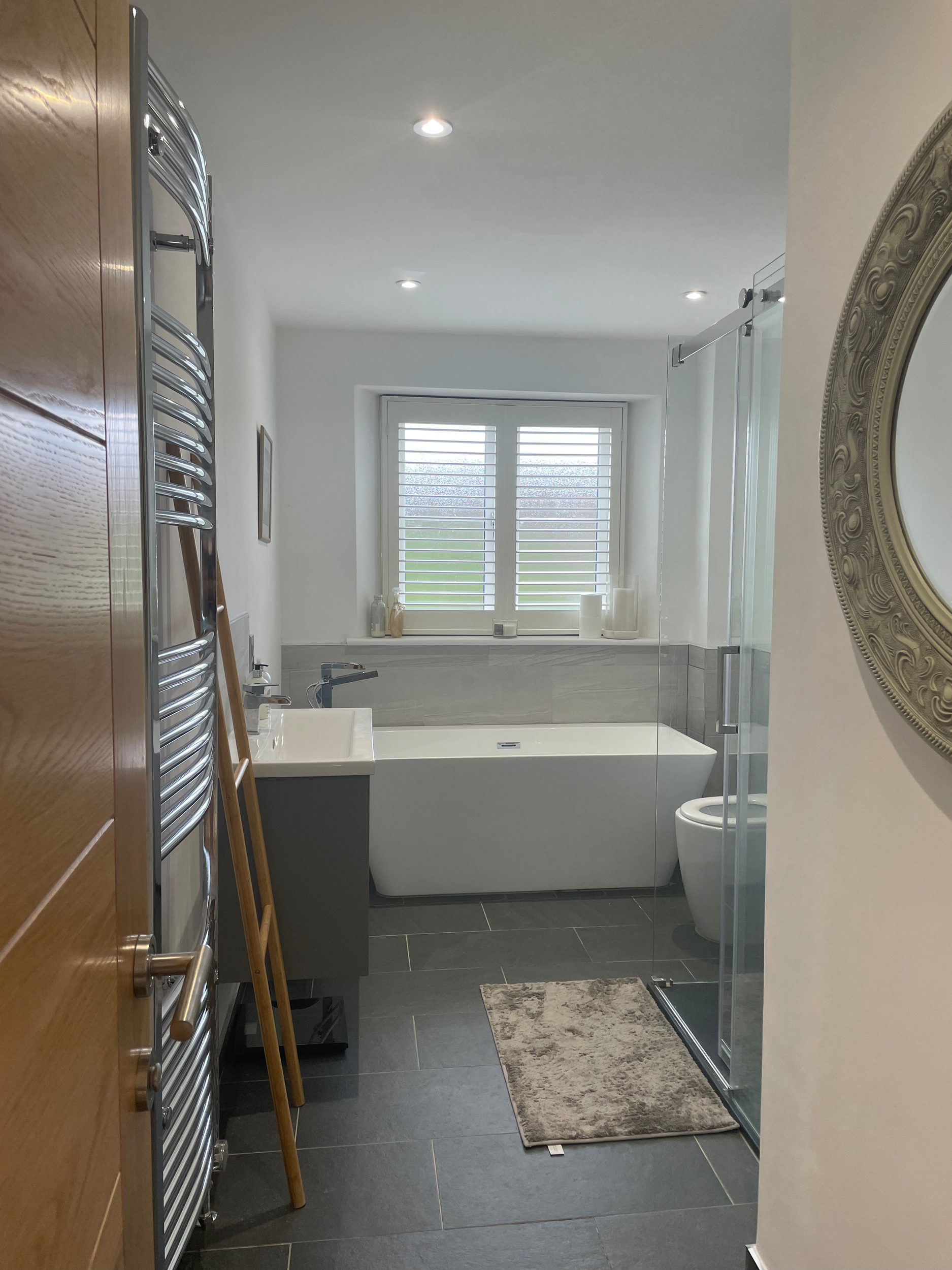 Modern bathroom with bathtub, toilet, window with shutters, gray tile floor, and a glass shower enclosure.