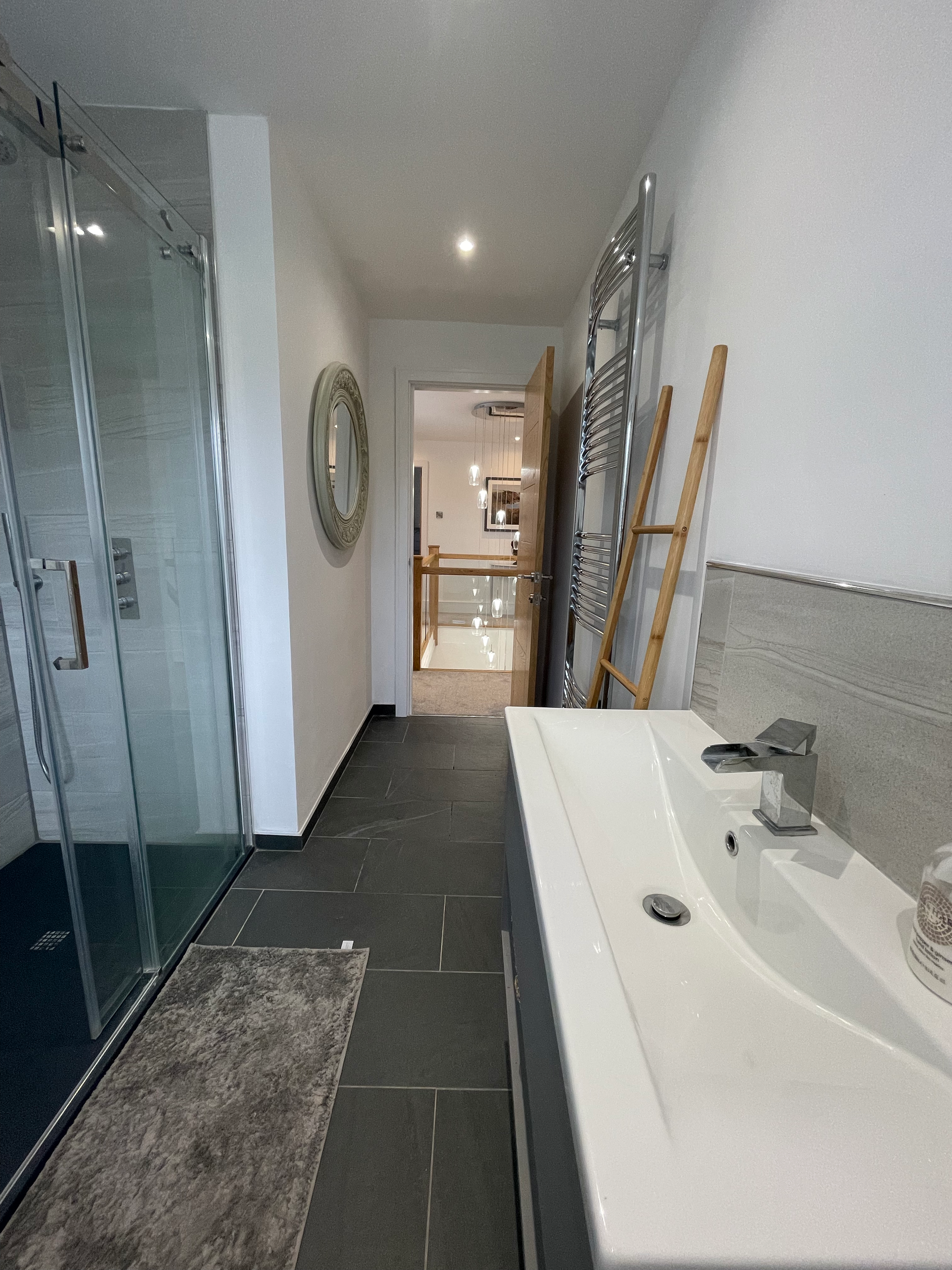 A modern bathroom with a glass shower, a white sink, a wall-mounted towel radiator, and a wooden ladder. The bathroom has dark floor tiles and a large mirror, with a door leading to a hallway.