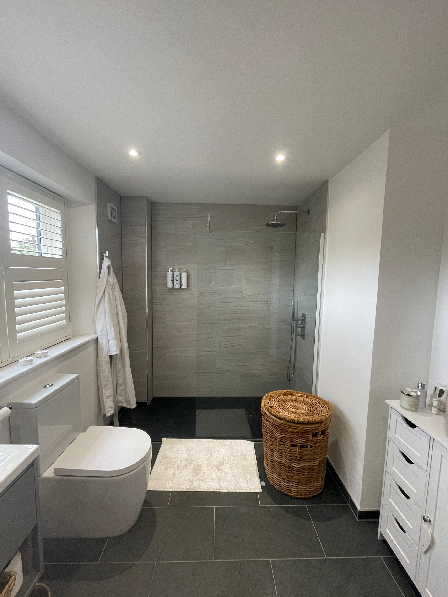 Modern bathroom with walk-in shower, white toilet, white cabinet, wicker laundry basket, and robe hanging on the wall near a window.