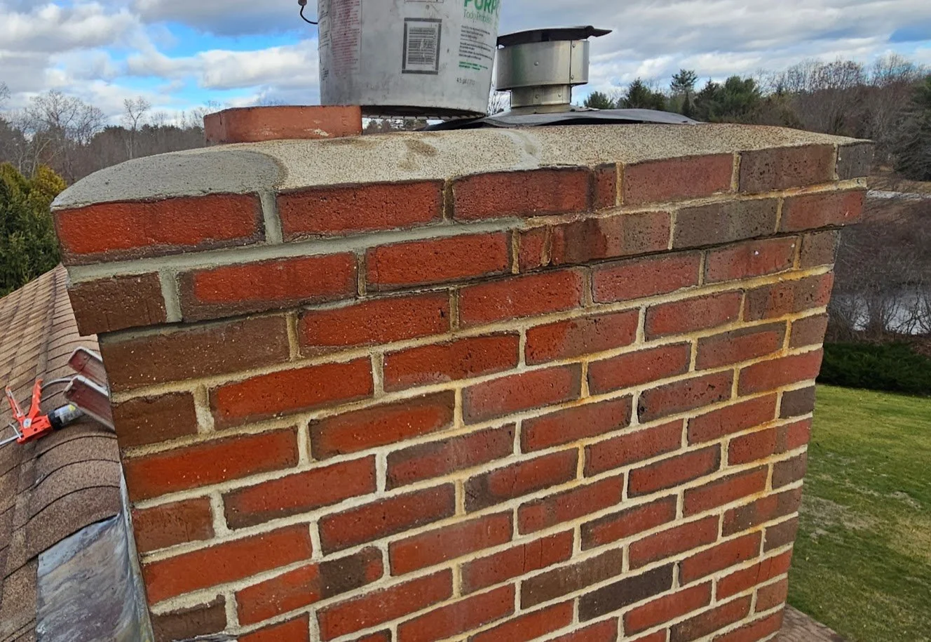 Close-up view of a brick chimney top with a metal cap, set against a partly cloudy sky and landscape in the background.
