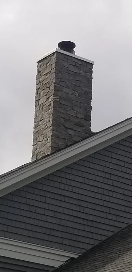 Close-up of a stone chimney on a house with dark gray siding and a sloped roof against an overcast sky.