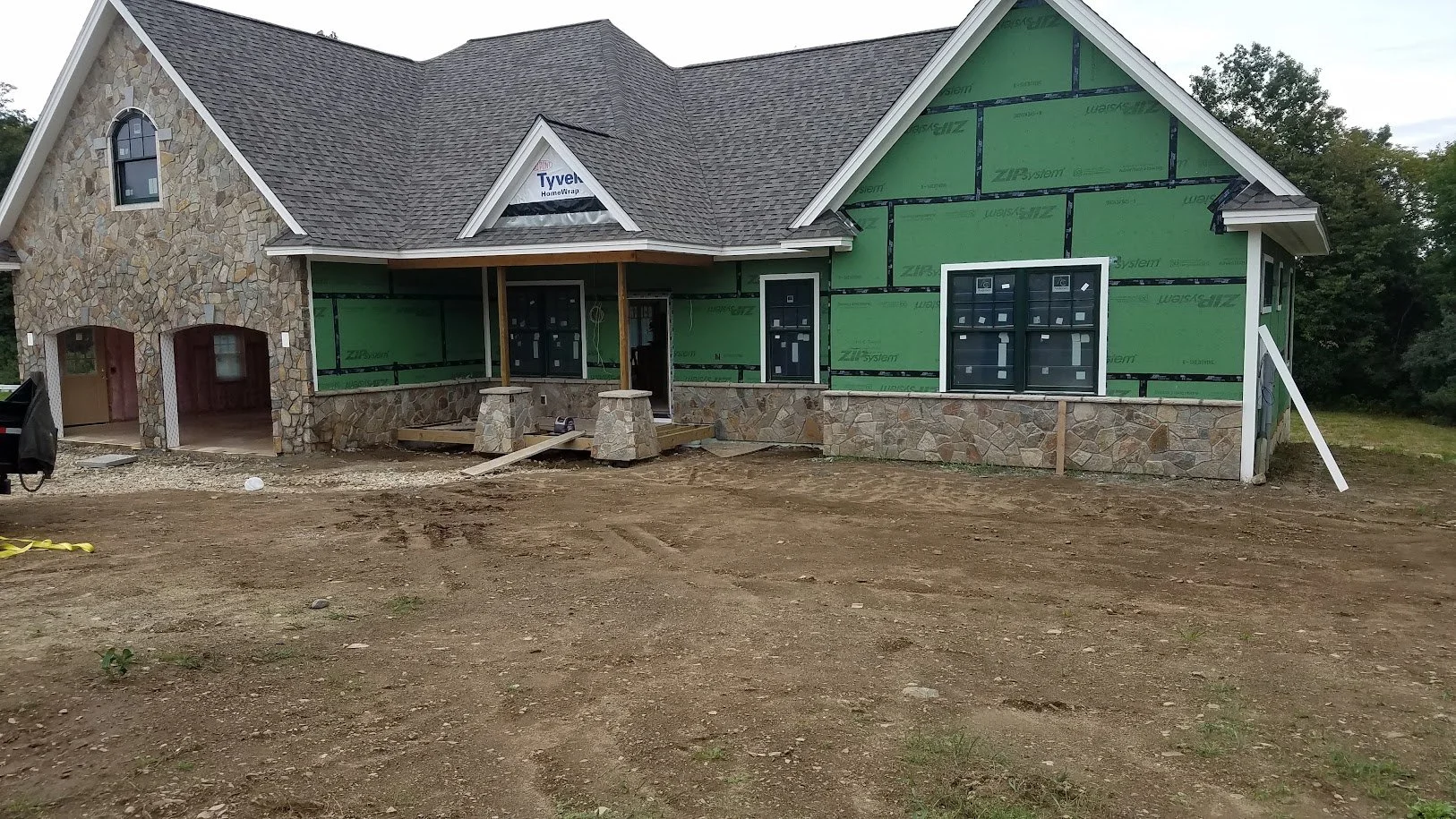 Under construction house with stone accents, green sheathing on part of the exterior, and a shingled roof. The front porch is unfinished, and the yard is dirt.