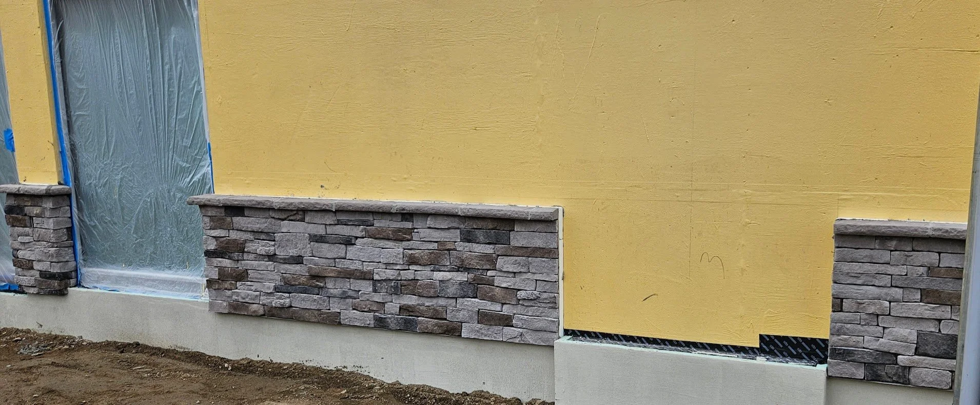 Construction site with a yellow wall, stone veneer accents, and wrapped window on the left, ground dirt in foreground.