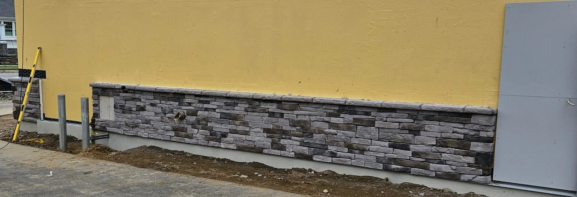 Construction site with a yellow wall, stonework trim, and gray electrical panel, with construction tools and dirt in the foreground.
