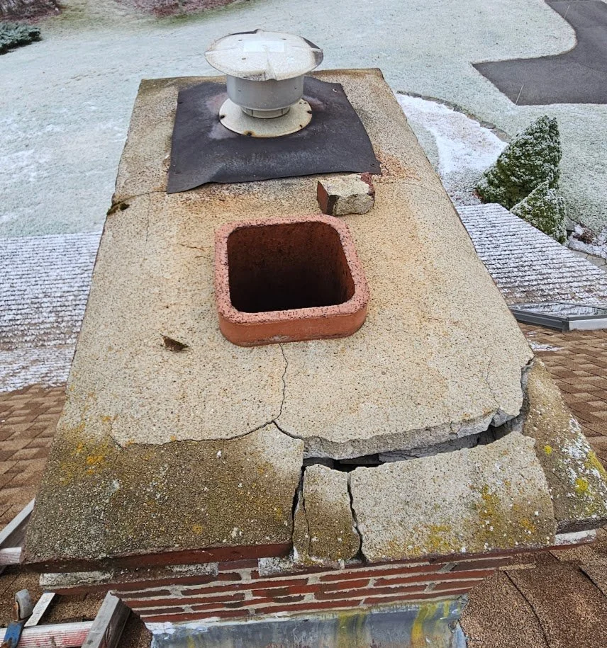 A cracked and weathered stone chimney with a round metal chimney cap on top, a small brick mortar, and an empty rectangular firebox opening.