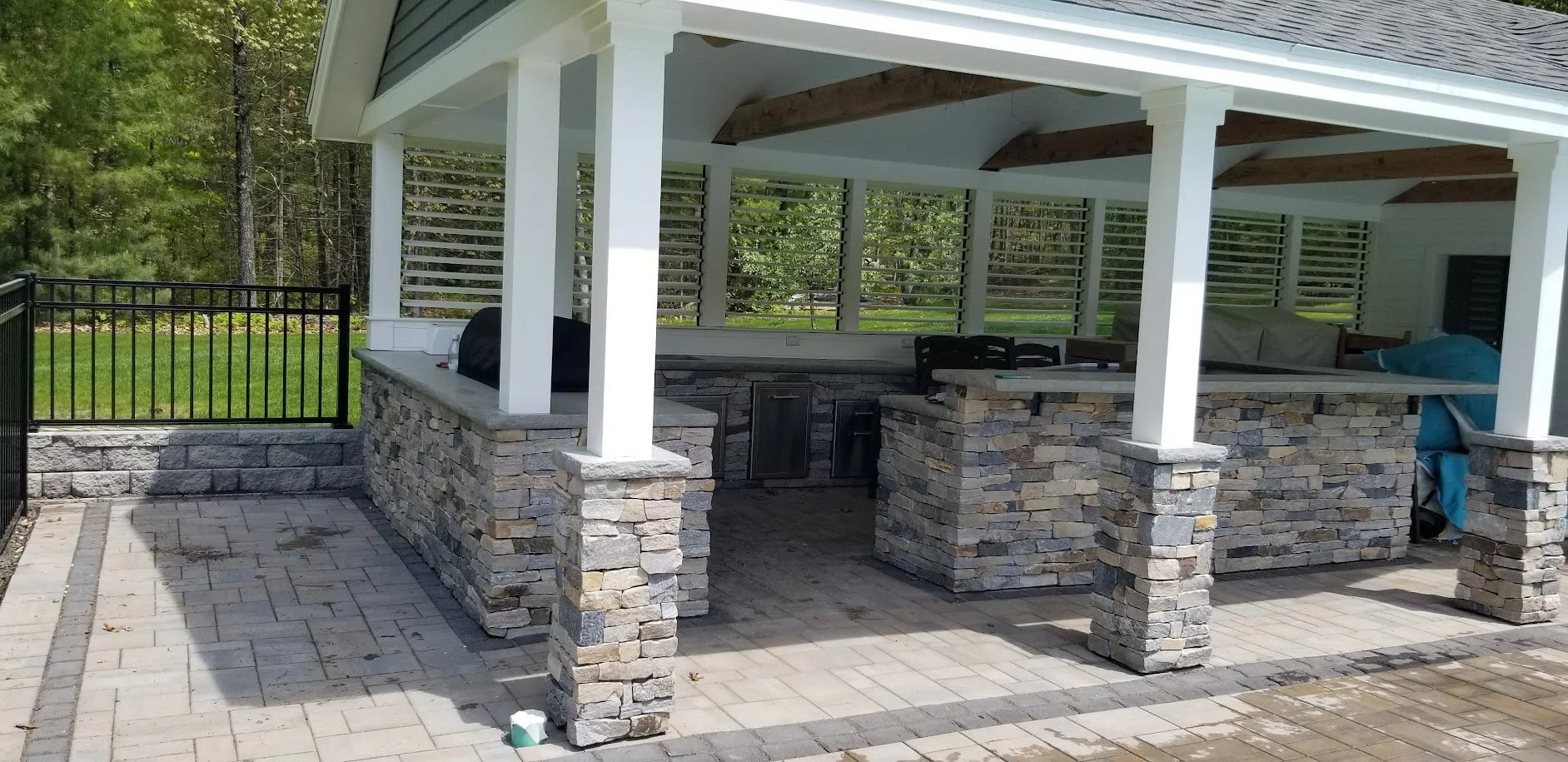 Finished outdoor patio with a stone bar and seating area, surrounded by a black metal fence and lush green trees in the background.