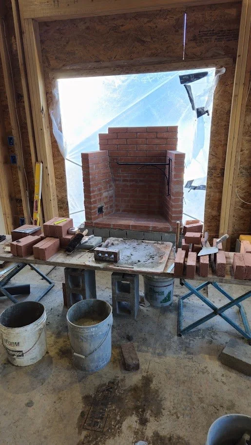 Construction site with a brick fireplace under construction inside a framed room, with building materials and tools scattered around.