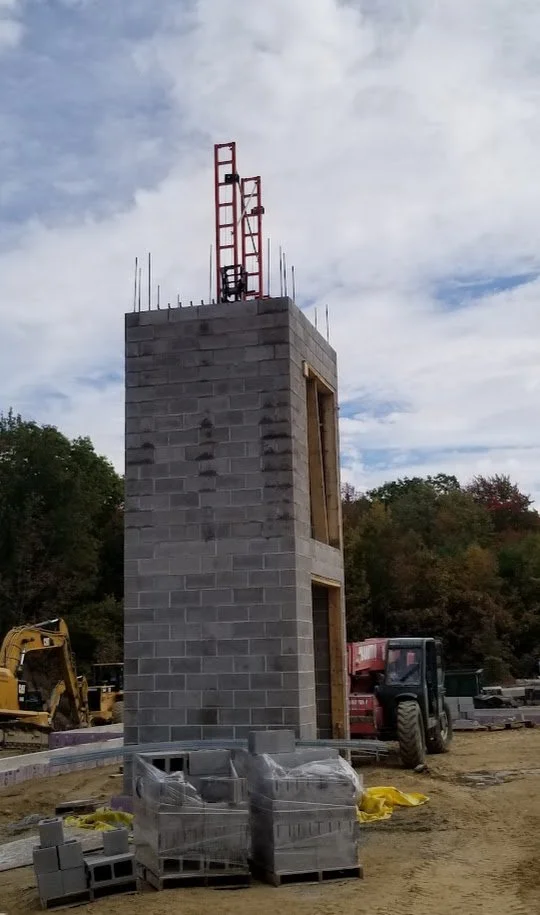 Construction site featuring a tall gray cinder block tower with window openings, construction equipment, and a cloudy sky.