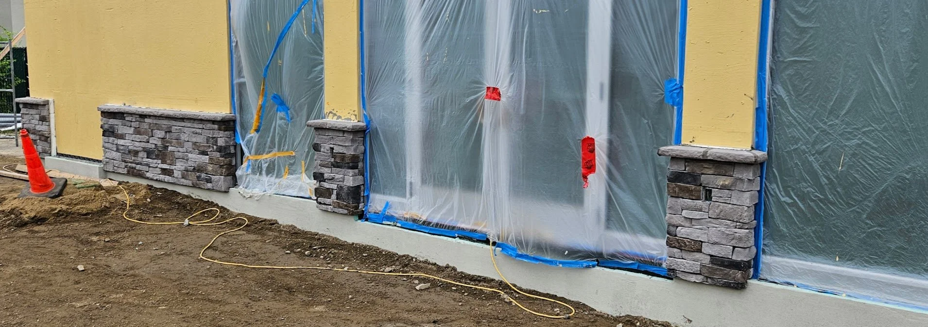 Front of a house under construction with yellow exterior walls, stone pillars, and plastic-covered windows and doors, with orange construction cone and electrical cords on the ground.