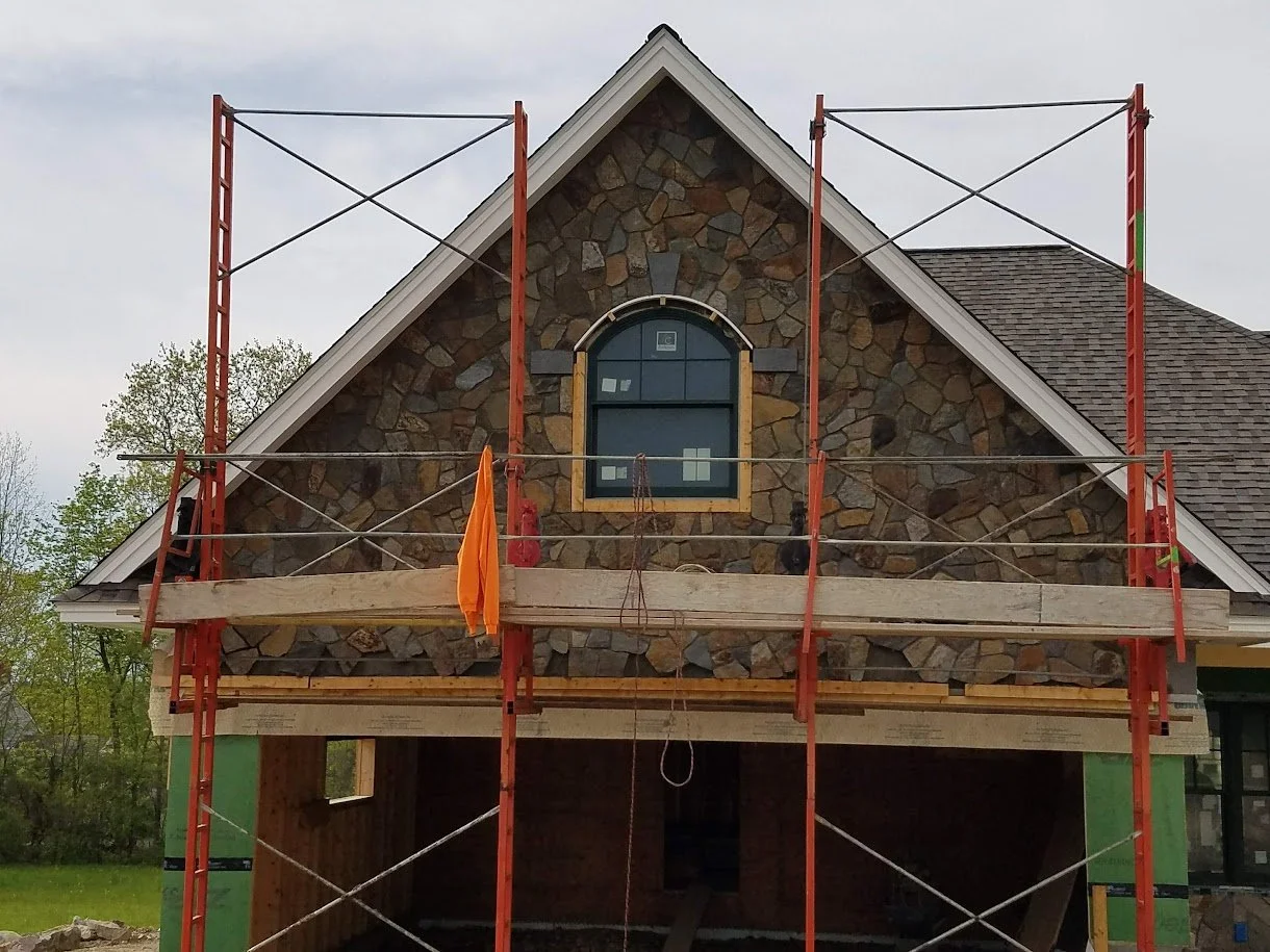 Construction scaffolding set up on a house with a stone facade and a window, indicating ongoing renovation or construction work.