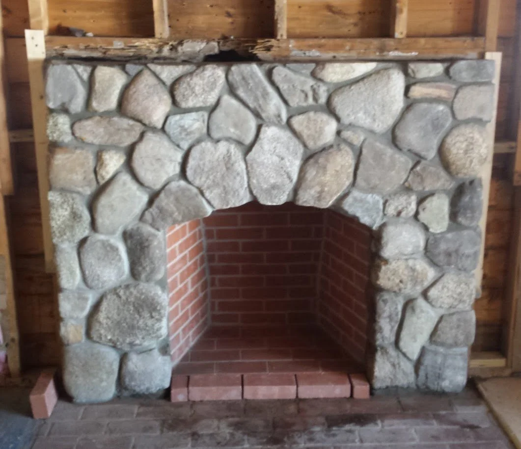 Stone fireplace with brick interior and wooden mantel in a construction site.