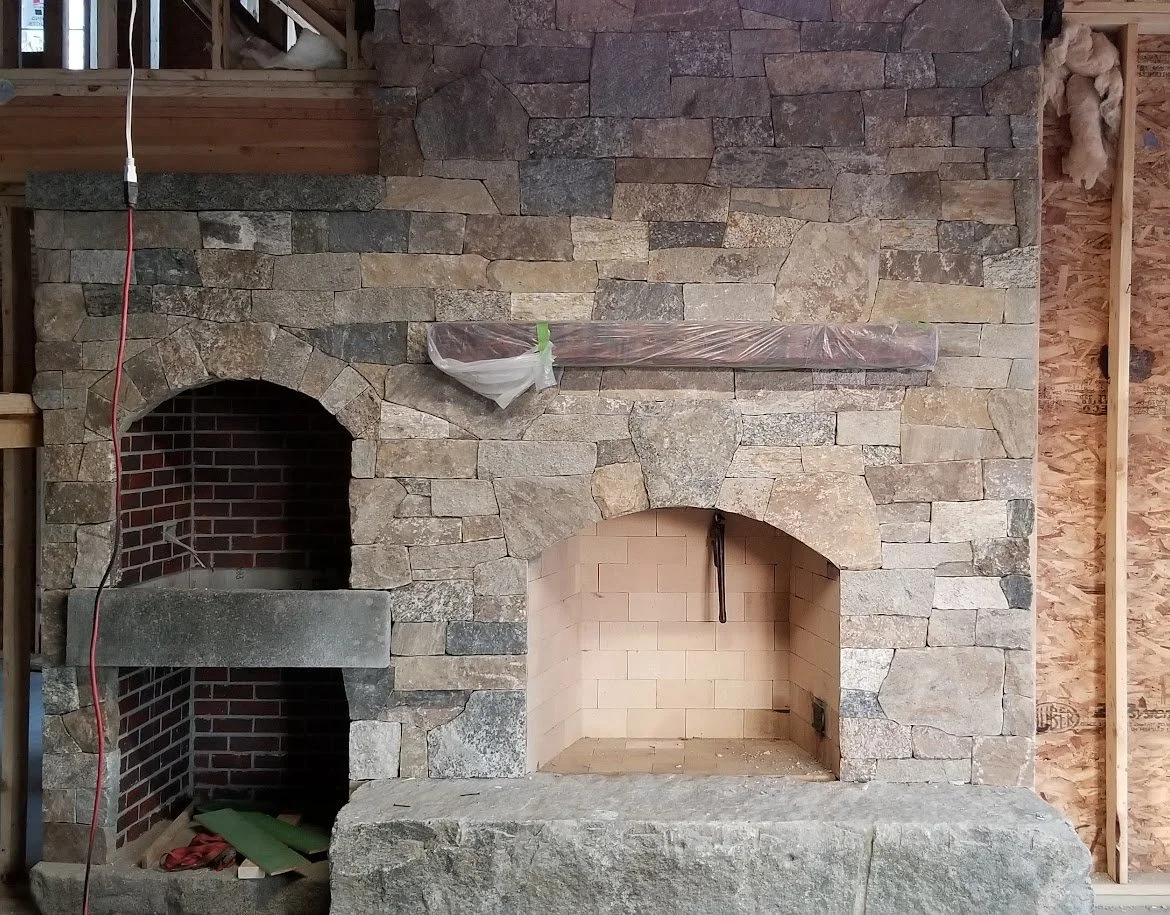 A stone fireplace under construction with a large stone hearth and two arched openings, one with brick interior on the left and the other with a lighter brick interior and tools hanging inside on the right.