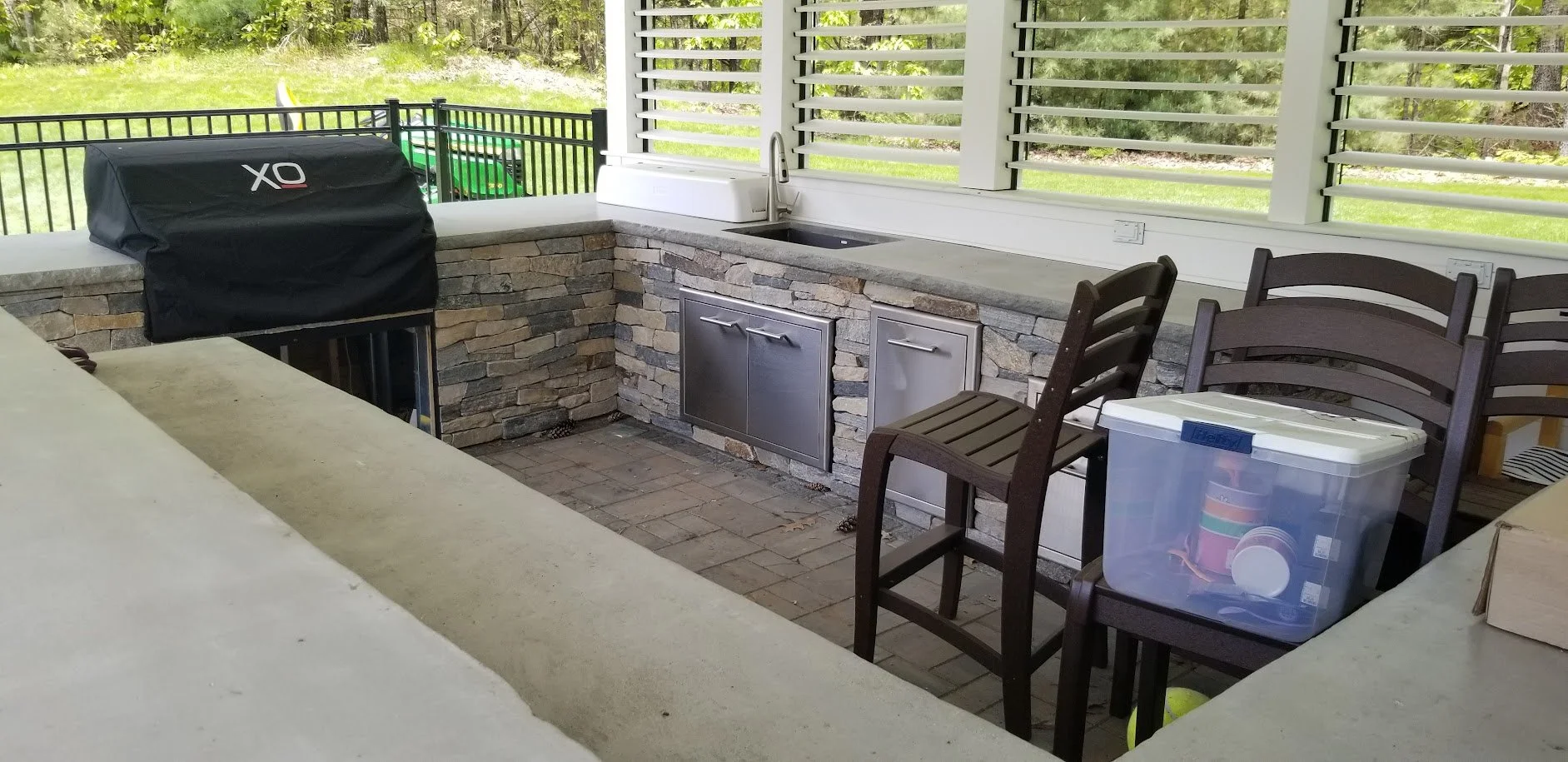 Outdoor kitchen area with a stone countertop, stainless steel sink, chairs, a plastic storage container, and a gas grill covered with a black protective cover. Green yard visible in the background.