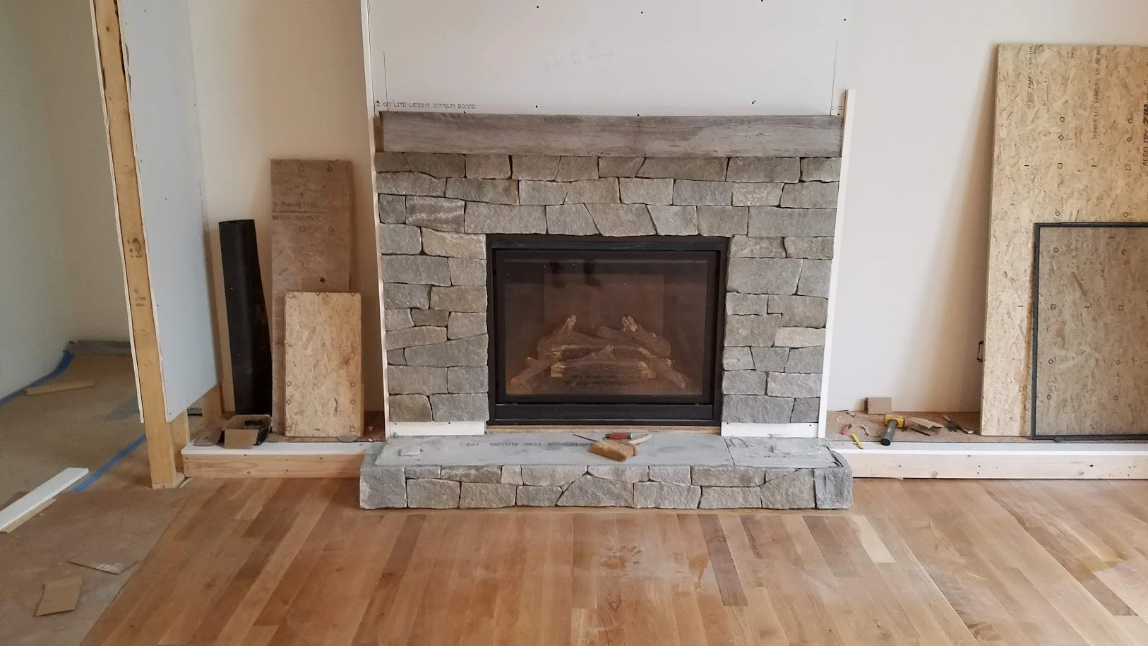 Living room fireplace surrounded by unfinished construction materials, with ledgestone facade, a concrete hearth, and a partially installed wooden mantel.