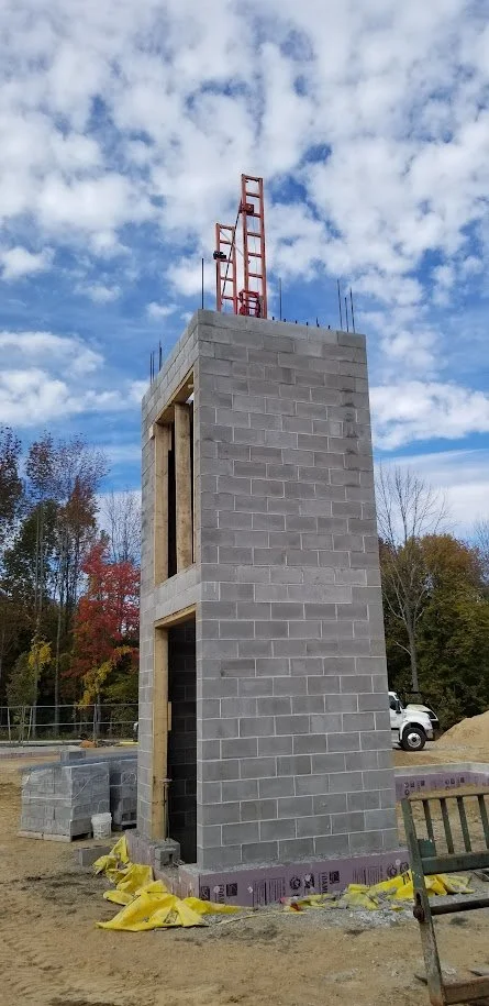Construction site with a gray cinder block tower under construction, with red scaffolding on top, and a partly cloudy sky in the background.