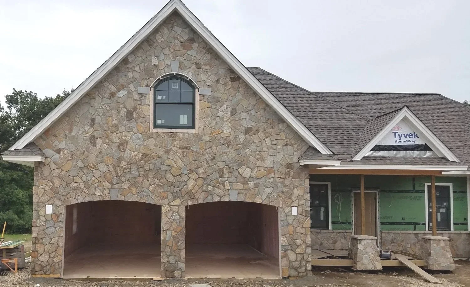Under construction house with a stone exterior, double garage, and partially completed porch, with an upper window and roofing shingles.