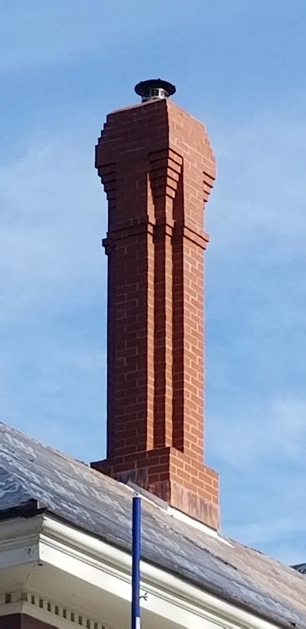 Close-up of a tall brick chimney with decorative brickwork, on a building roof under a clear blue sky.
