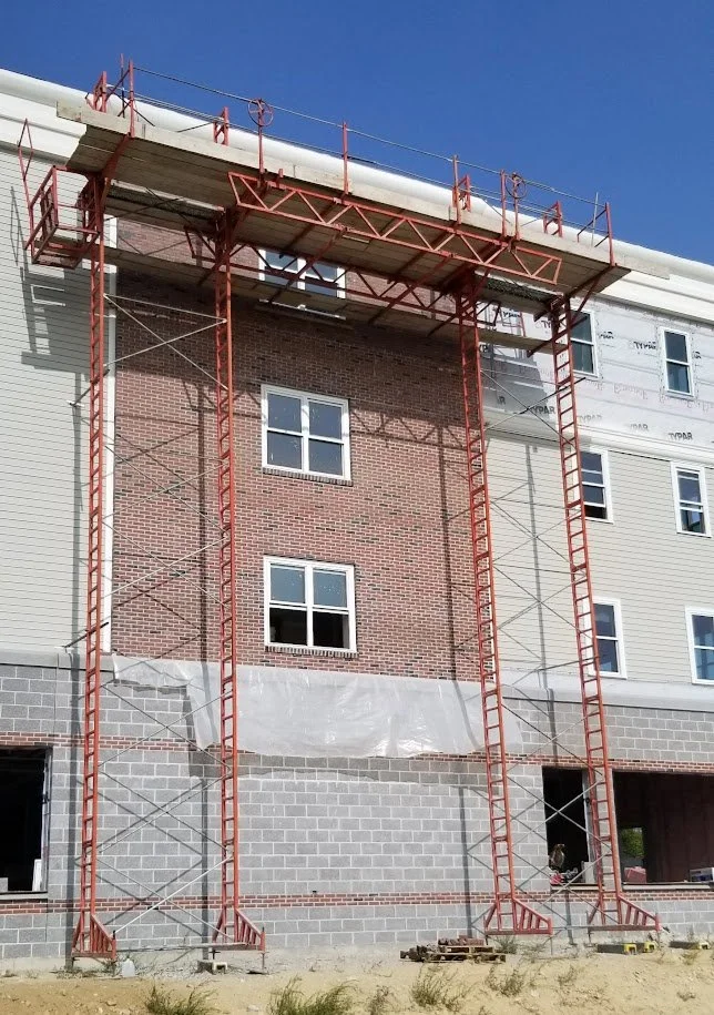 Construction scaffolding around the exterior of a multi-story building, with brick and siding walls, and open window spaces.
