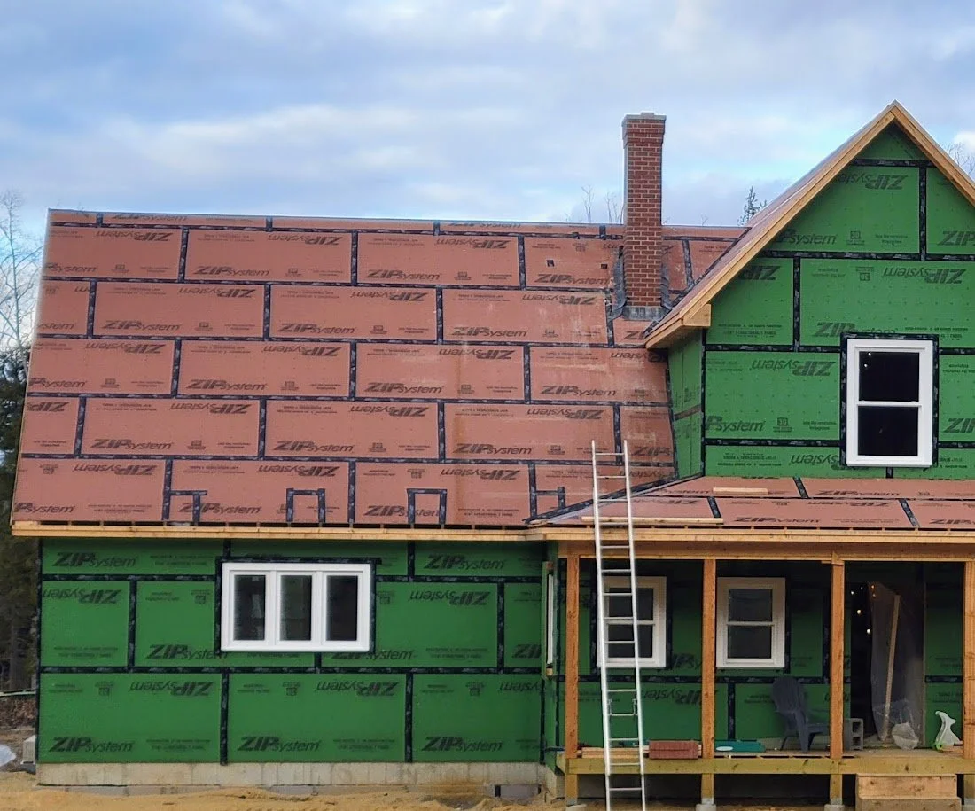 House under construction with green and orange building wrap, a ladder leaning on it, and a brick chimney on the roof.