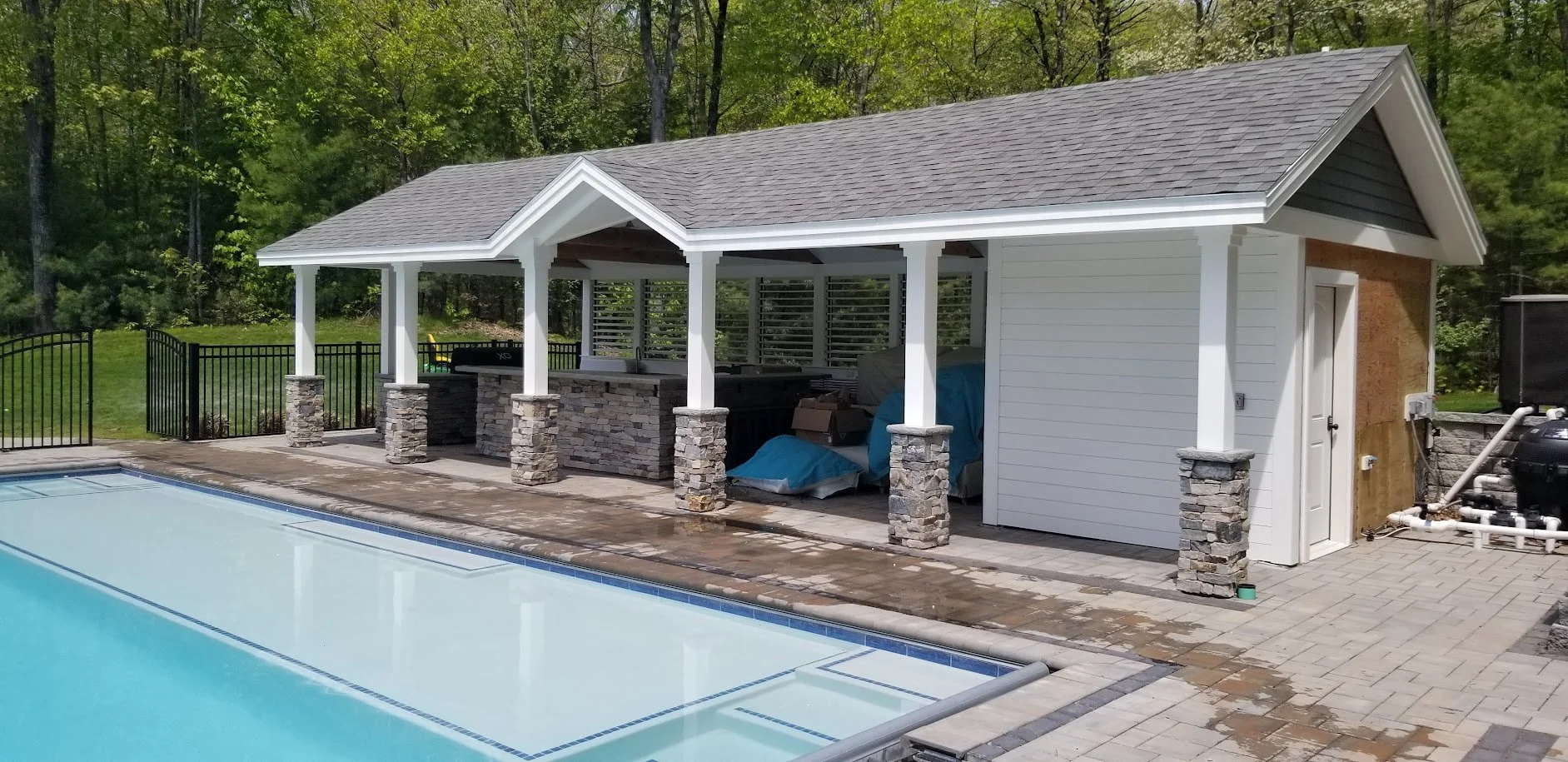 Backyard pool area with a covered outdoor kitchen and storage shed, surrounded by a forested area with green trees.