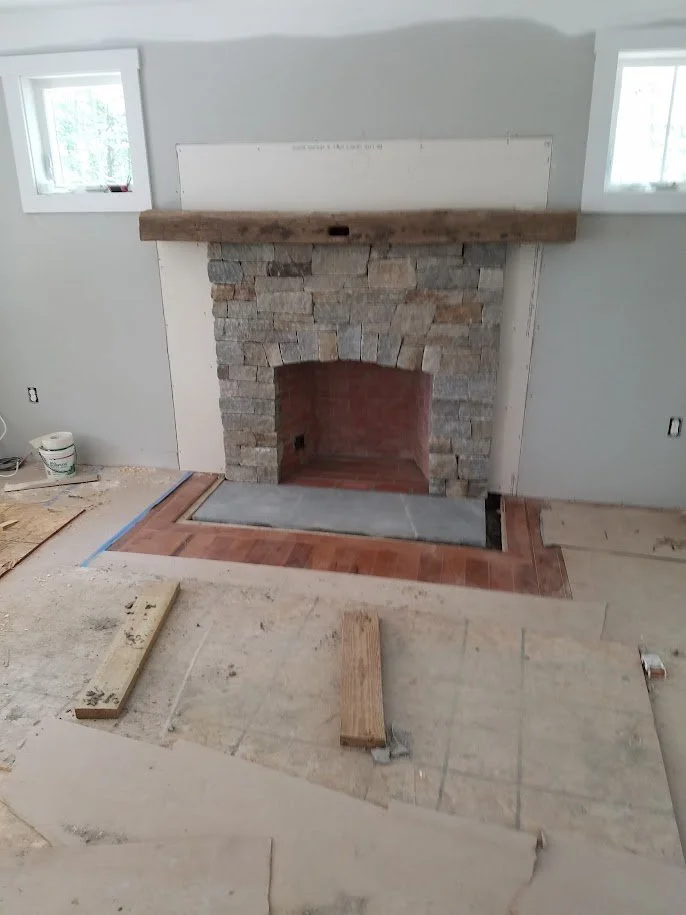 Indoor fireplace under construction with stone surround and wooden mantle, with new flooring work in progress.