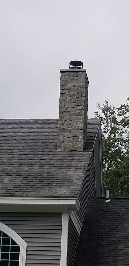 A stone chimney on the roof of a house with gray siding and a gray shingle roof, overcast sky in the background.