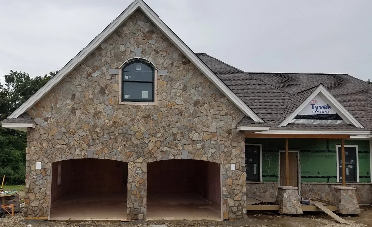 Side view of a house under construction with stone exterior, two garage open bays, a window, and a front porch with stone pillars