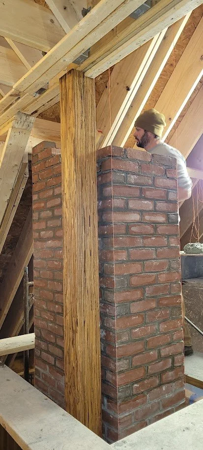 A man working on a brick chimney in an attic, with exposed wooden beams and support structures around him.