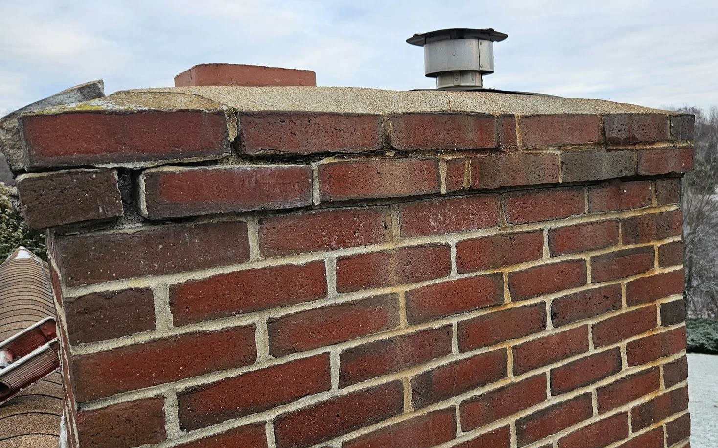 Close-up of a brick chimney with metal vent on top, part of roof visible on the left, and cloudy sky and trees in the background.