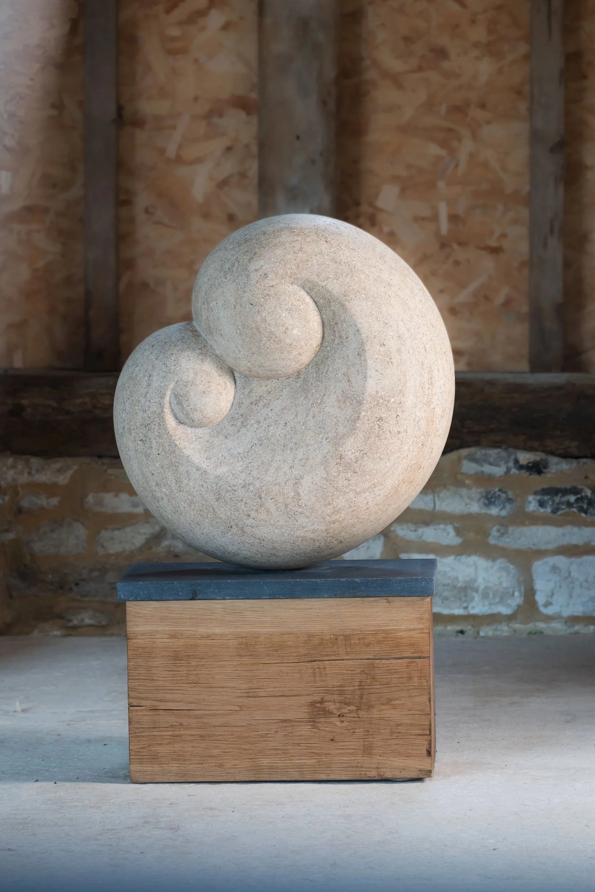 A spiral-shaped stone sculpture made by Dominic Welch with two spherical protrusions, mounted on a wooden plinth in front of a rustic wood and brick background.