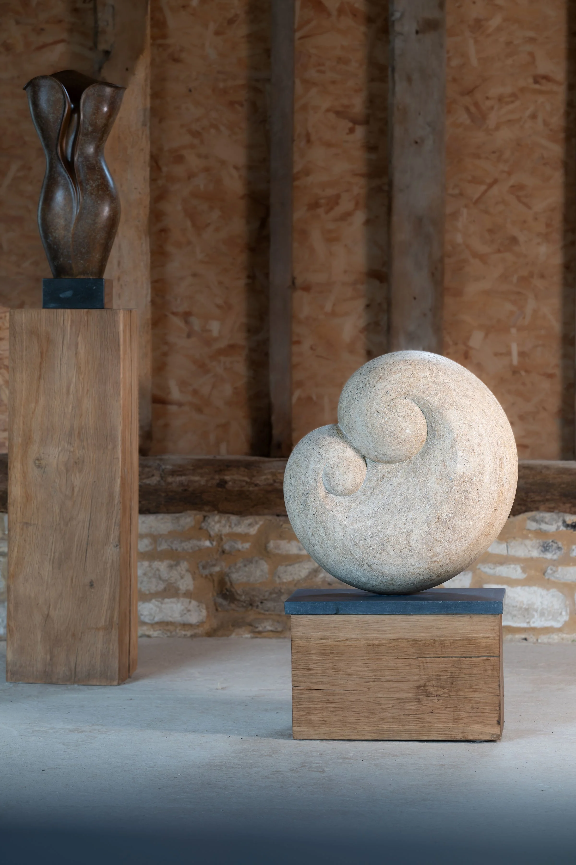 Two abstract sculptures, one tall made of bronze on a wooden pedestal, and one spiral-shaped stone on a wooden base, inside a rustic room with wooden walls.
