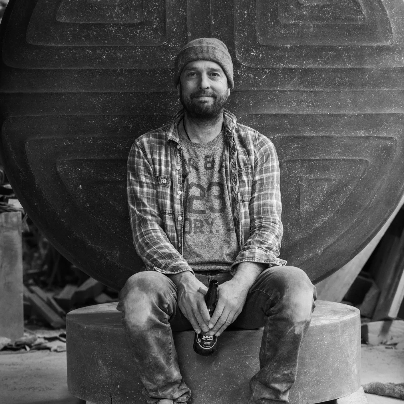 A man with a beard and a beanie sitting on a large stone, holding a bottle of beer, in a workshop with a large, rounded object behind him.