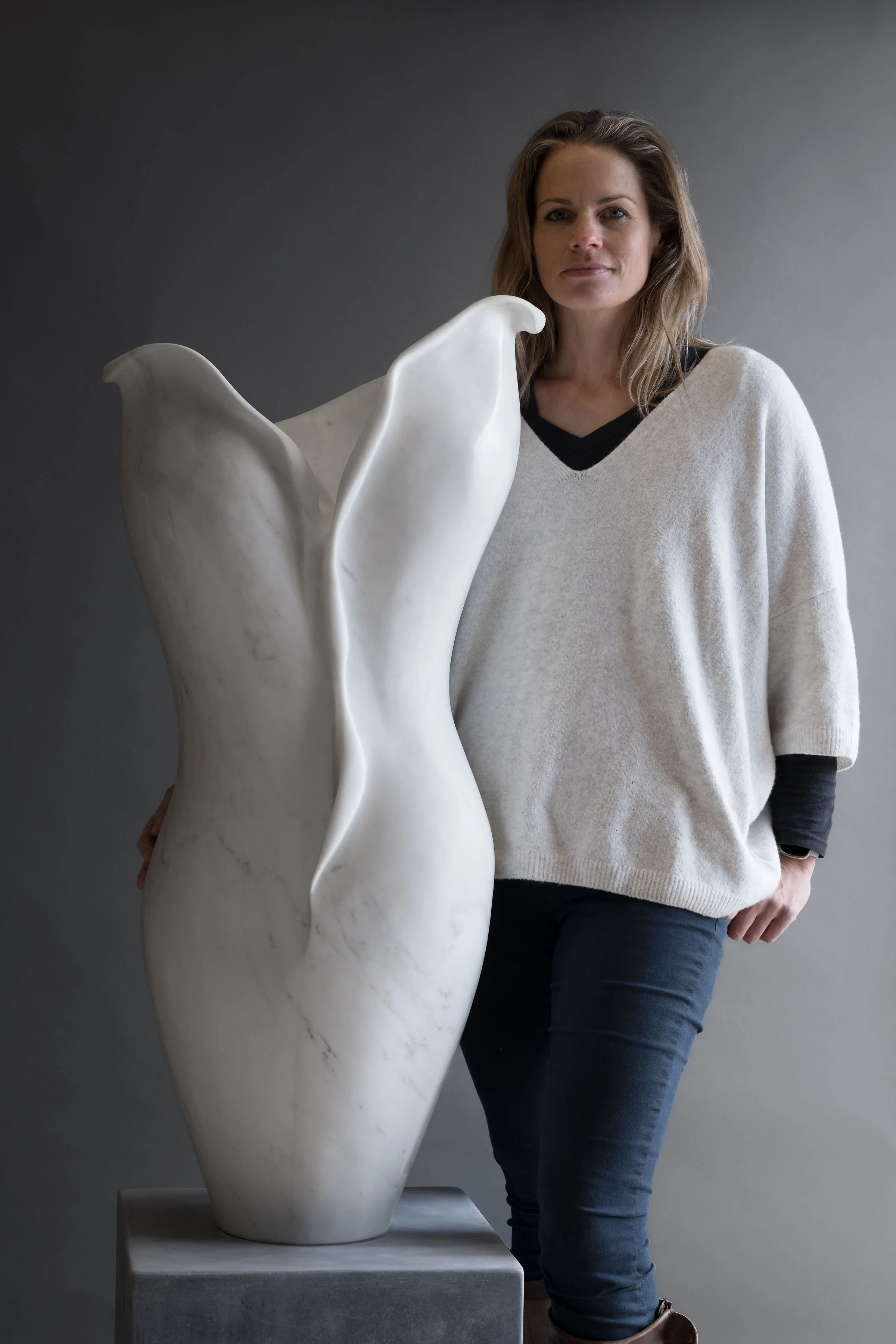 A woman standing next to a large abstract white sculpture of torso, against a gray background.