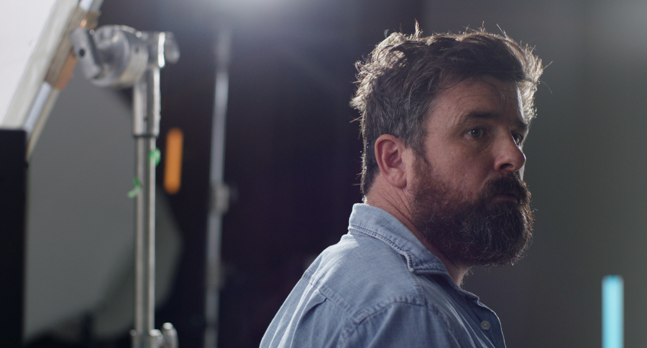 A man with a beard and messy hair looking to the side in a studio or photo shoot setting.